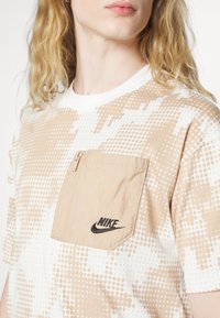 Kortärmad, beige och vit mönstrad T-shirt med en dragkedjeficka. Har en svart Nike-logotyp på fickan. Tillverkad av bomulls material.