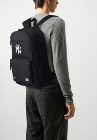 Schwarzer Stoffrucksack mit aufgesticktem weißem Logo der New York Yankees, mit Reißverschlussfächern, verstellbaren Trägern und MLB-Label an der Unterseite.