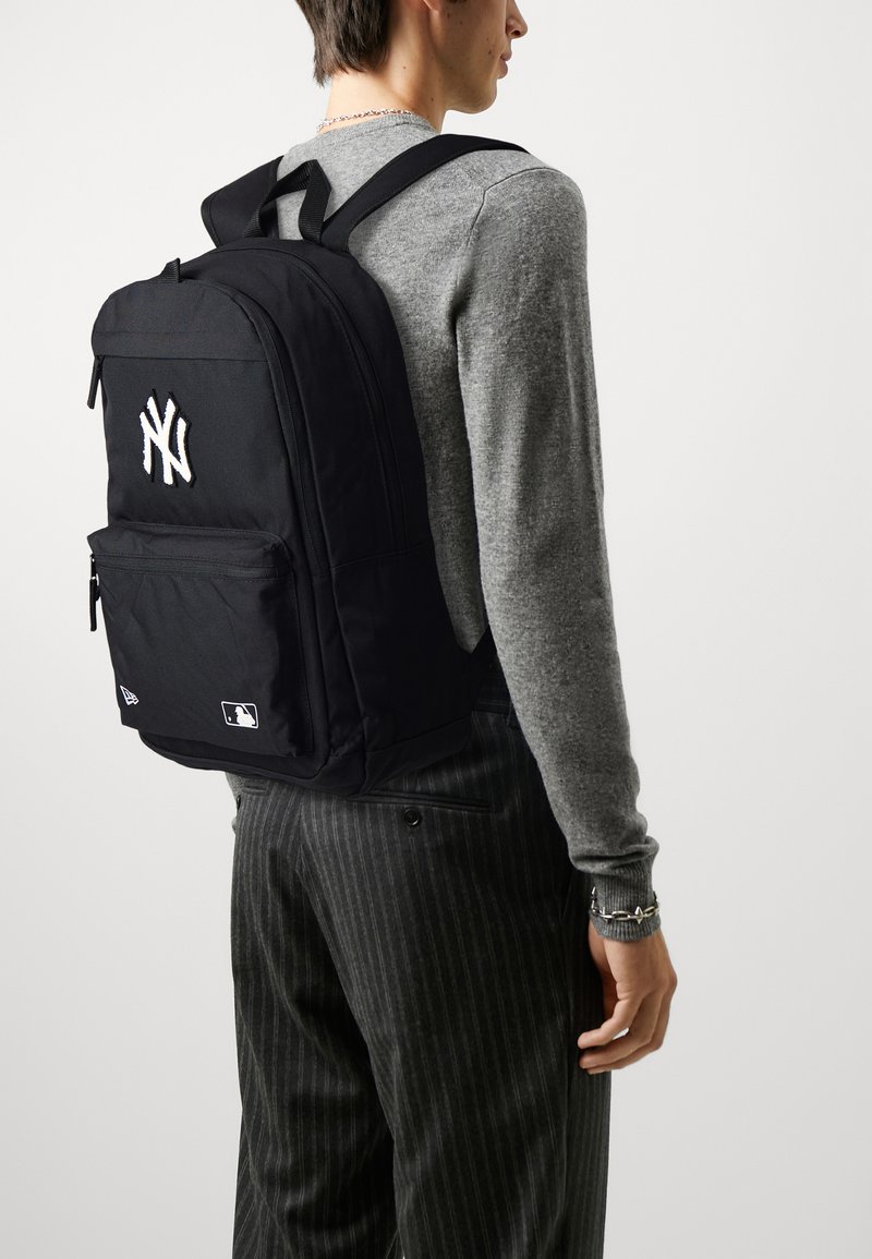 Schwarzer Stoffrucksack mit aufgesticktem weißem Logo der New York Yankees, mit Reißverschlussfächern, verstellbaren Trägern und MLB-Label an der Unterseite.