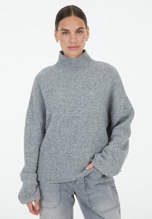 Grauer Strickpullover mit hohem Stehkragen, der eine strukturierte Oberfläche und kleine, dekorative Perlenakzente entlang des Saums und der Ärmel aufweist.