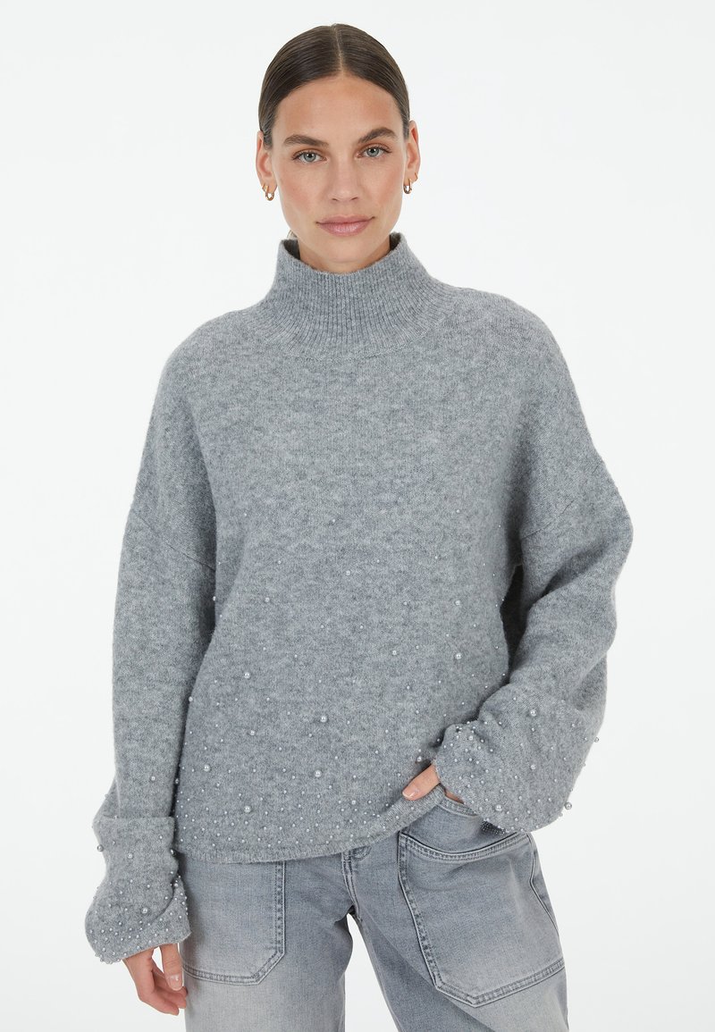 Grauer Strickpullover mit hohem Stehkragen, der eine strukturierte Oberfläche und kleine, dekorative Perlenakzente entlang des Saums und der Ärmel aufweist.