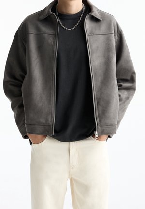 Person wearing a gray zippered jacket over a black shirt and light beige pants with hands in pockets, shown from shoulders to hips.