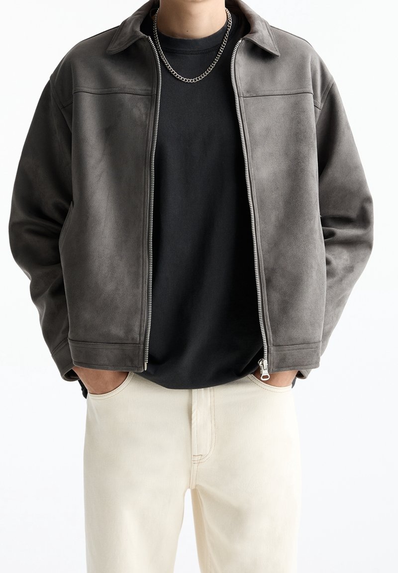 Personne portant une veste grise zippée par-dessus une chemise noire et un pantalon beige clair, les mains dans les poches, montrée des épaules jusqu'aux hanches.