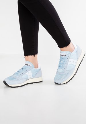 Persoon die lichtblauwe Saucony-sneakers en zwarte skinny broek draagt, staand op een witte vloer met één voet iets opgeheven.