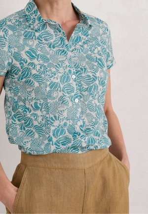 Personne portant une chemise à manches courtes boutonnée avec des motifs coquillages bleus et blancs, rentrée dans un pantalon marron clair taille haute.