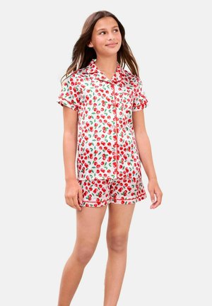 Mädchen steht, trägt ein weißes kurzärmeliges Hemd mit Knöpfen und passende Shorts mit rotem Kirschmuster und grünen Blättern.