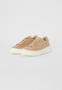 Beige suede sneakers with a chunky white sole, featuring flat laces and side stitching details. Rounded toe design and minimalistic profile.