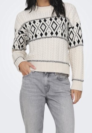 Frau trägt einen cremefarbenen Pullover mit Zopfmuster und schwarzen geometrischen Mustern auf Brust und Ärmeln, kombiniert mit hellgrauen Jeans.