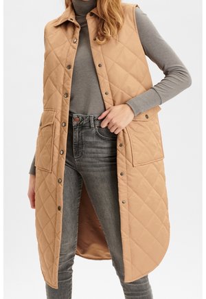 Femme portant une longue doudoune beige sur un col roulé gris et un jean gris, ajustant la doudoune près de la taille avec la main.