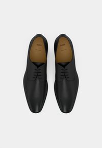 Une paire de chaussures de cérémonie en cuir noir verni avec des lacets, semelles intérieures marquées "BOSS Fabriqué en Italie", vues de dessus sur fond blanc.
