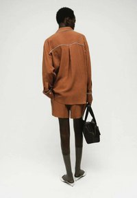 Persona in piedi con una camicia e pantaloni corti color ruggine, che tiene in mano una borsa nera, indossando collant trasparenti scuri e tacchi bianchi a punta su uno sfondo semplice.
