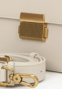 Bílá kožená kabelka se zlatě pruhovanou kovovou sponou. Obsahuje odpovídající bílý popruh s kovovým klipem ve zlaté barvě. Hladký povrch.