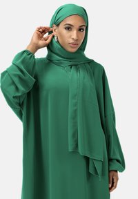 Grön, flödande lång klänning med matchande hijab. Tyget ser mjukt ut med en svag lyster. Har lösa ärmar och en draperad sjaldetalj.