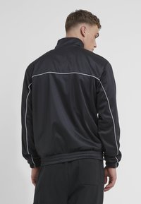 Black athletic jacket with a high collar, white piping along the shoulders and back, and ribbed hem and cuffs. Smooth, lightweight fabric.