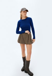 Top a maniche lunghe blu a coste abbinato a una gonna houndstooth marrone. Stivali marroni fino al ginocchio con dettaglio in fibbia. Un berretto grigio completa l'outfit.