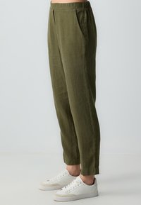 Olivegrüne Leinenhose mit lockerer Passform, ausgestattet mit Seitentaschen und elastischem Bund, kombiniert mit weißen Sneakers. Geschmeidige Textur.
