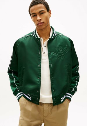 Młody mężczyzna w ciemnozielonej kurtce varsity z białymi paskami i haftowanymi inicjałami, białej koszulce polo oraz beżowych spodniach.