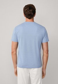 Mies, jolla on lyhyet ruskeat hiukset, pukeutuneena vaaleansinaiseen lyhythihaiseen T-paitaan ja valkoisiin housuihin, seisoo selkä kameraan päin yksinkertaista taustaa vasten.