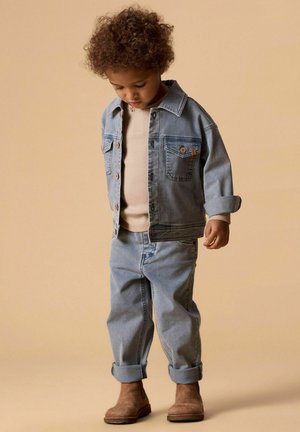 Käharate juustega väikelaps, kes kannab helerohelist teksajope ja teksapükse, beežkat särki ja pruune saapaid, seisab ja vaatab alla.