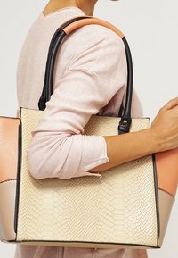 Tote de cuero sintético color marfil con estampado de pitón, asas negras y acentos en melocotón, que presenta un diseño estructurado y una textura suave.