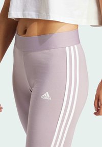 Lila leggingi iz raztegljivega materiala, s visokim pasom in tremi belimi črtami ob straneh ter majhnim logotipom na stegnu.