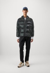Donsjack in donkergroen, met een glanzende afwerking, twee grote vo zakken en een ritssluiting. Gedragen over een gestreept shirt en zwarte broek.