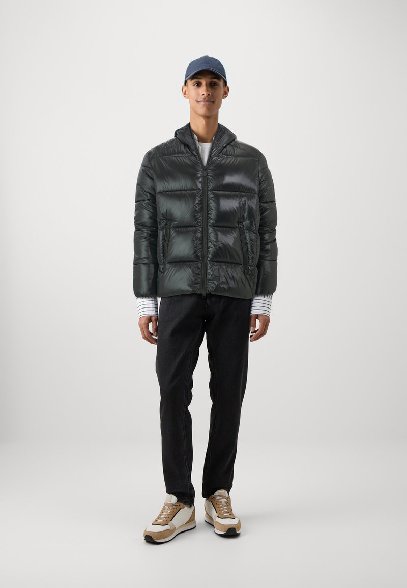 Giacca a vento verde scuro, con una finitura lucida, due grandi tasche frontali e chiusura con zip. Indossata sopra una maglietta a righe e pantaloni neri.