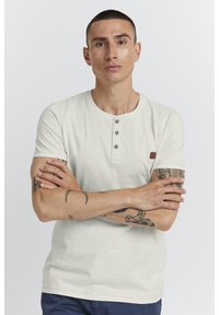 Fehér, rövid ujjú póló, három gombos gombolással és egy barna márkajelzés a bal vállnál. Simább pamut textúra.