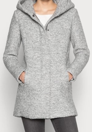 Femme portant un manteau à capuche texturé gris clair avec poches latérales, associé à un pantalon gris foncé et un haut blanc en dessous.