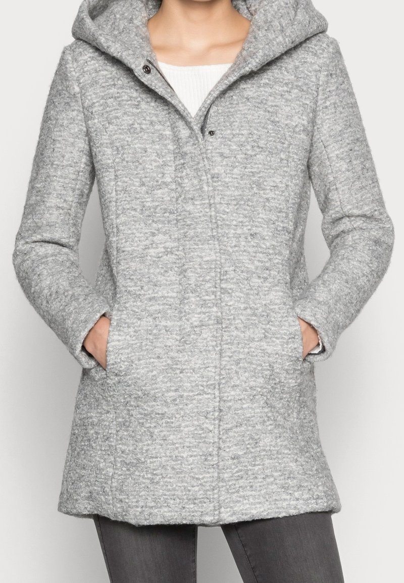 Femme portant un manteau à capuche texturé gris clair avec poches latérales, associé à un pantalon gris foncé et un haut blanc en dessous.