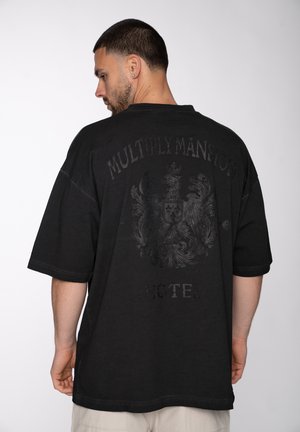 Man with short hair and beard wearing oversized black t-shirt with dark crest and "Multiply Mansion Hotel" text, facing away.