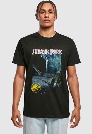 Personen iført svart Jurassic Park T-skjorte som viser dinosaurhode nær en annen person ved en bil, med Jurassic Park-tittelen og den gule dinosaurlogoen.