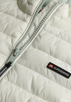 Gevoerde jas in off-white, met een grijze ritssluiting en een zwart label met rode accenten. Het materiaal heeft een gladde textuur en een doorgestikt ontwerp.