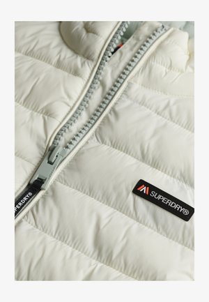 Veste matelassée de couleur blanc cassé, dotée d'une fermeture éclair grise et d'une étiquette noire avec des accents rouges. Le matériau présente une texture lisse et un design matelassé.