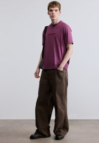 Μωβ μπλουζάκι με μαύρο λογότυπο, συνδυασμένο με καφέ παντελόνι με φαρδιά μπατζάκια. Το μοντέλο στέκεται μπροστά σε ουδέτερο φόντο, φορώντας μαύρα παπούτσια.