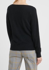 Pull en tricot noir avec des manches longues et un col large. Présente des poignets et un ourlet côtelés. Porté sur un pantalon à carreaux gris clair avec des accents jaunes.
