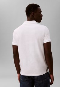 Fehér rövid ujjú póló klasszikus gallérral, strukturált anyagból, oldalsó hasítékokkal. Kék farmernadrággal viselve.