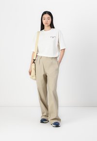 Pantalon beige ample à taille haute, associé à un t-shirt oversize blanc avec un logo noir, et des baskets bleu marine et blanches.