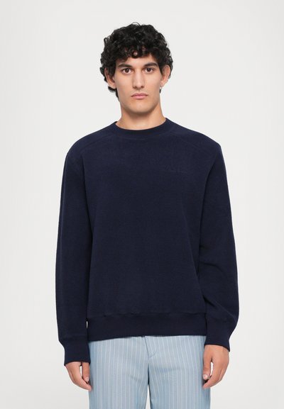 Mann mit lockigem Haar, der einen dunkelblauen Langarmpullover und eine hellblaue, gestreifte Hose trägt, und vor einem schlichten weißen Hintergrund steht.