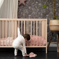 Trækradle med lodrette balustre, lys naturlig finish og en blød lyserød madras. Barn i hvidt sæt bøjer sig for at samle op lyserøde stofformer.