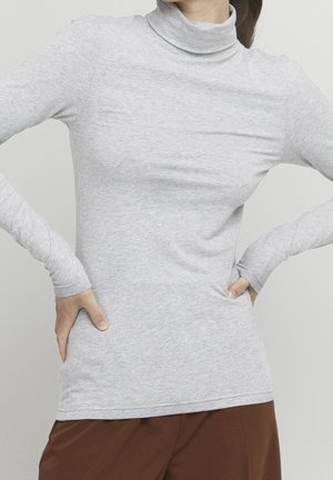 Personne portant un pull à col roulé à manches longues gris clair, les mains sur les hanches, associée à un pantalon marron, sur un fond uni.