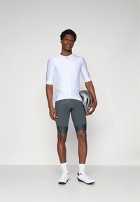 Mann i hvit kortermet sykkeltrøye, grå polstrede sykkelshorts, hvite sokker og sko, som holder en grå sykkelhjelm ved sin side.