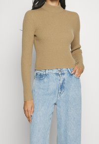 Jersey de cuello alto beige acanalado con mangas largas, diseño ajustado y textura suave, combinado con jeans de tiro alto en azul claro.