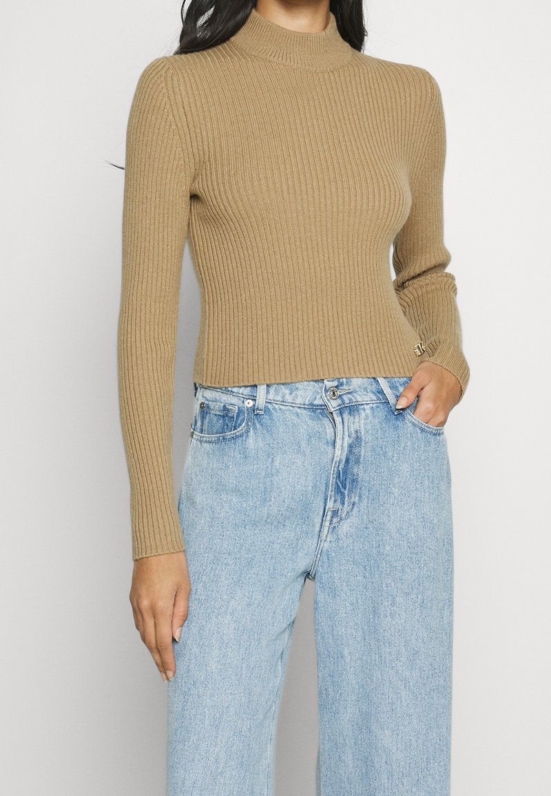 Jersey de cuello alto beige acanalado con mangas largas, diseño ajustado y textura suave, combinado con jeans de tiro alto en azul claro.