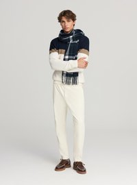 Maglione a maglia con righe blu navy, beige e bianche, abbinato a una sciarpa plaid oversize nei toni del blu navy e verde. Pantaloni color crema e scarpe marroni.