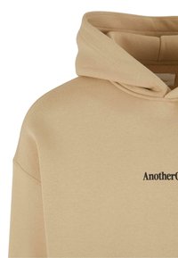 Beige hoodie met een zachte textuur, voorzien van een grote capuchon en een voorzak. De merknaam is in het zwart op de borst gedrukt.