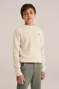 Gaiši bežs ribots džemperis ar apaļo kaklu un nelielu izsistu logotipu. Apvienots ar olīvu zaļiem teksturētiem biksēm, parādot ikdienišķu fitu.