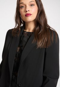 Blazer nero dal design strutturato, con chiusura a un solo bottone e tessuto liscio. Sotto, una maglia trasparente con dettagli piumati.