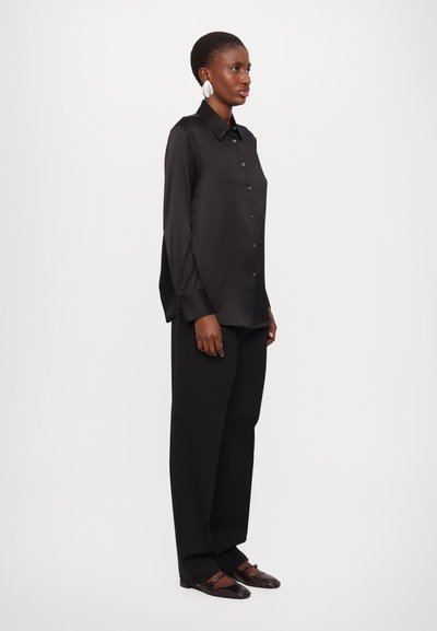 Chemise noire à manches longues avec col, associée à un pantalon noir ample et des chaussures marron à motifs. Texture de tissu lisse.