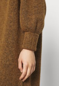 Brun, fluffig stickad klänning med detalj som har en lång, lös ärm med ribbad manschett, vilket framhäver textur och värme. Handen placerad nära sömmen.